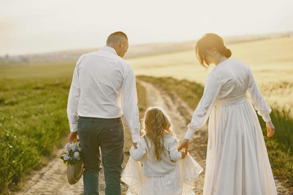A young couple walking down a path holding both hands of a young girl in white on a sunny day. Symbolizing God-given identity and joy
