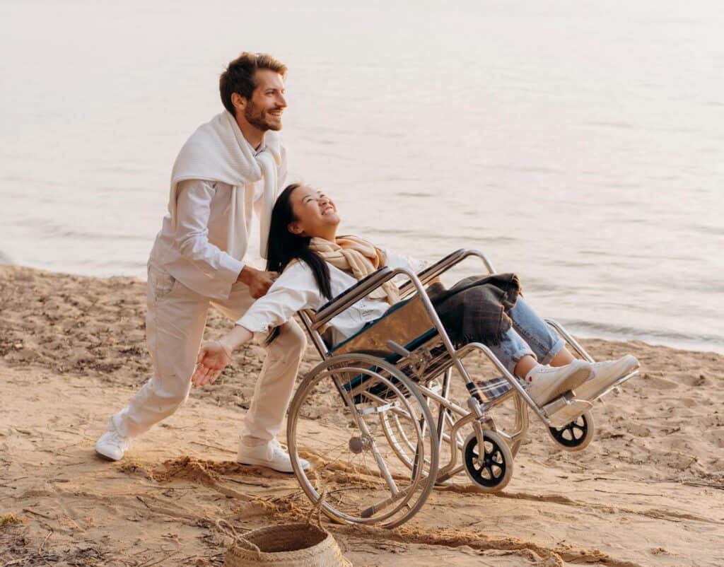 a man pushing a woman in a wheelchair on the beech as she smiles symbolizing love in action as service