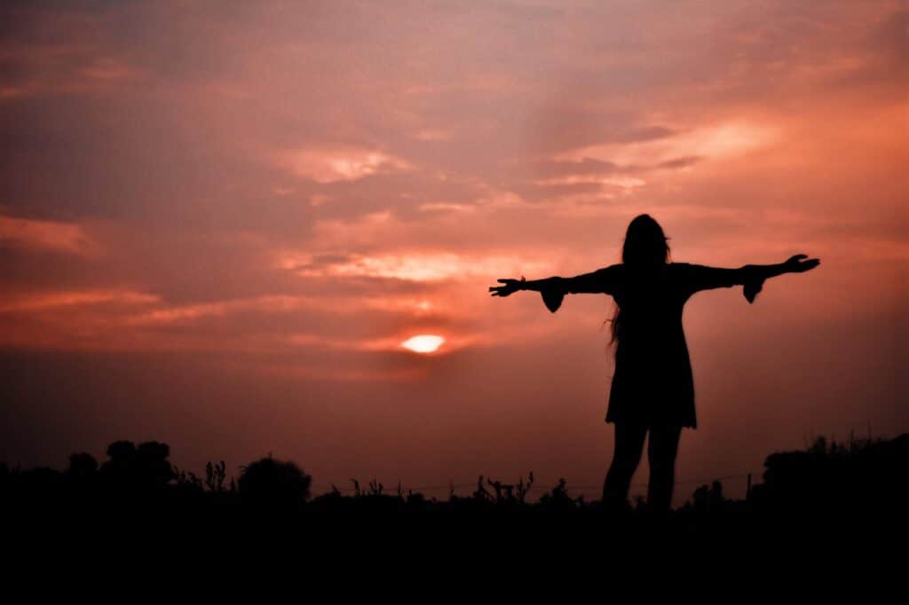 a woman standing in a field at sunset with her arms outstretched towards the sky seeking God's Love